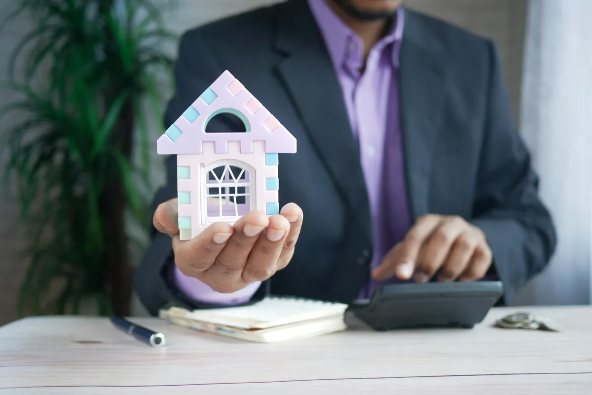 Person holding a house model beside a calculator